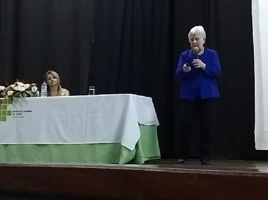 Professora Mara Rúbia sentada e professora Mirza em pé, de casaco azul escuro e calça preta. Senhora de estatura baixa e cabelos brancos